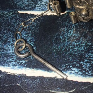 May include: Close-up of a silver-toned metal clasp with a chain and key-shaped component. The clasp has decorative floral elements and coiled springs. The key-shaped piece is connected to the chain by a small ring.