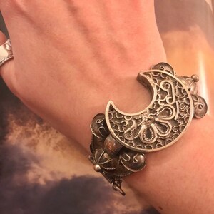 May include: Silver-toned cuff bracelet featuring a crescent moon design with intricate filigree and floral details. The bracelet has a hinged closure and is adorned with additional decorative elements. The jewelry is worn on the wrist.