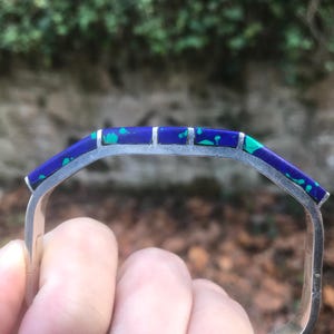 May include: A silver-toned bracelet with a rectangular design. The top of the bracelet features a vibrant blue inlay with green accents. The bracelet is held by a hand, with a blurred background of green foliage.