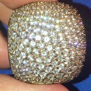 May include: A square-shaped ring covered in numerous small, sparkling clear stones. The ring has a gold-colored band and a surface completely encrusted with the glittering stones, creating a dazzling effect. The ring is held against a blue background.