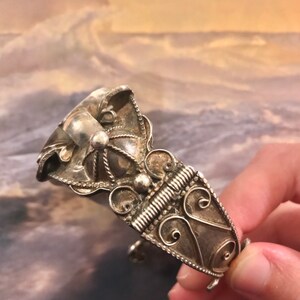 May include: Ornate silver-toned cuff bracelet featuring intricate filigree and beaded details. The bracelet has a hinged design with a decorative clasp. The design includes swirling patterns and a central, raised element.