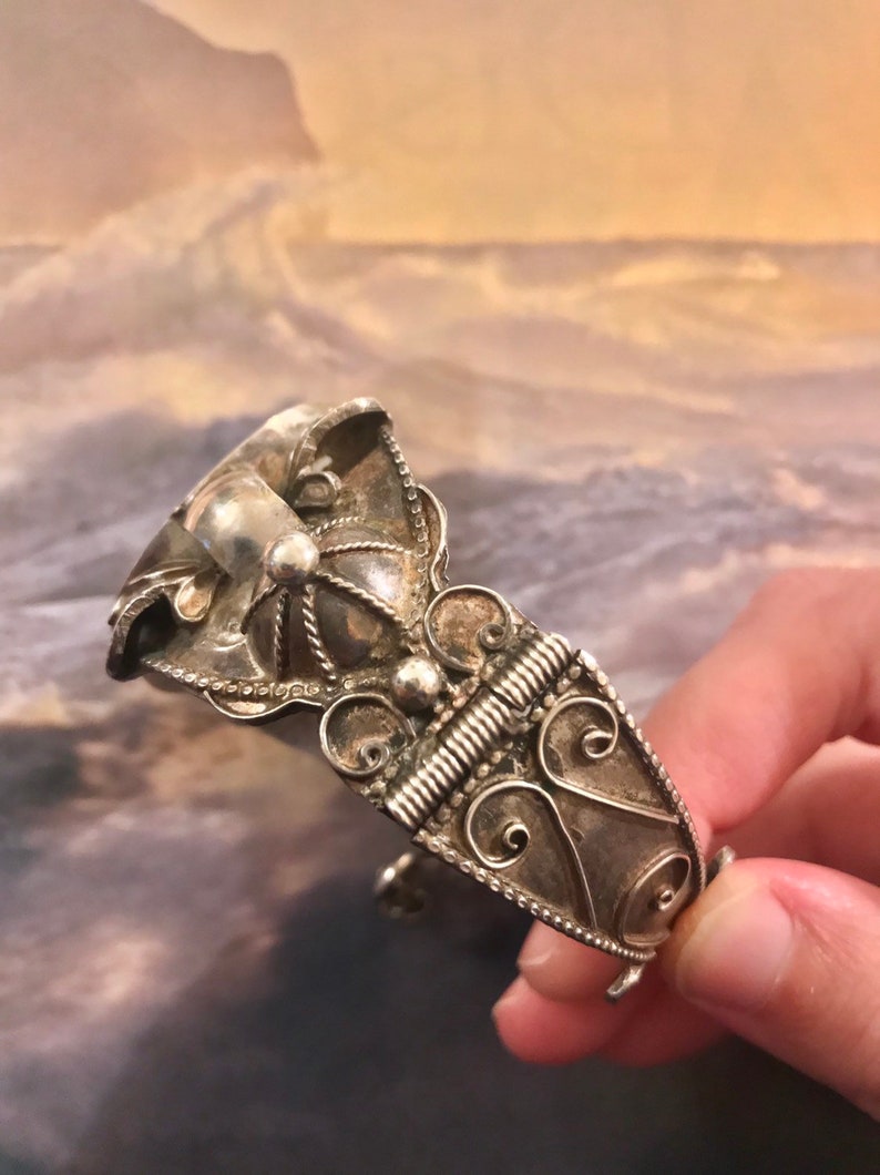 May include: Ornate silver-toned cuff bracelet featuring intricate filigree and beaded details. The bracelet has a hinged design with a decorative clasp. The design includes swirling patterns and a central, raised element.