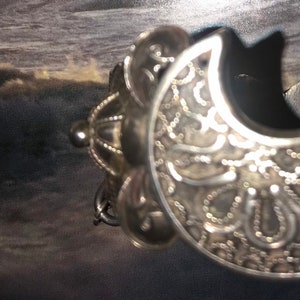 May include: Ornate silver-toned metal bracelet with a crescent shape. The bracelet features intricate filigree and beaded detailing. The design includes a decorative element with a small, round bead and a chain.