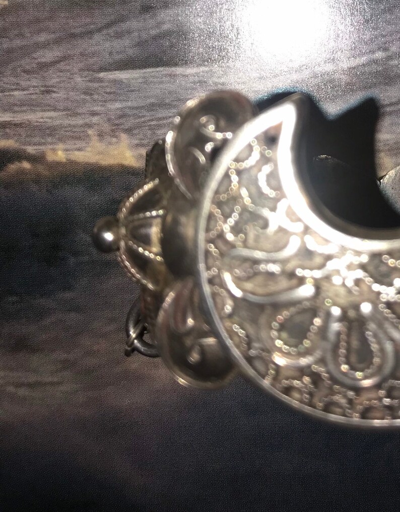 May include: Ornate silver-toned metal bracelet with a crescent shape. The bracelet features intricate filigree and beaded detailing. The design includes a decorative element with a small, round bead and a chain.