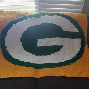 May include: A handmade, crocheted blanket in the colors of the Green Bay Packers. The blanket features a large, white "G" logo with a green outline on a yellow background. The blanket is folded over a couch.
