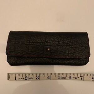 May include: A black leather wallet with a red stitch detail and a small metal button. The wallet is closed and lying flat on a white surface.