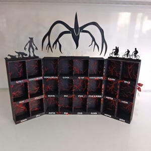 Könnte beinhalten: Eine schwarze Holzvitrine mit mehreren Fächern, die rote, venenartige Muster aufweist. Die Vitrine ist mit Silhouetten von Charakteren und Kreaturen verziert, und die Namen der Charaktere sind aufgedruckt.