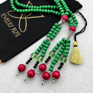 May include: Green wooden prayer beads with red and black accents, silver metal accents, and a yellow tassel. The beads are strung on a black cord. The beads are displayed on a black fabric pouch with the text "DZN JEWELRY" in gold lettering.