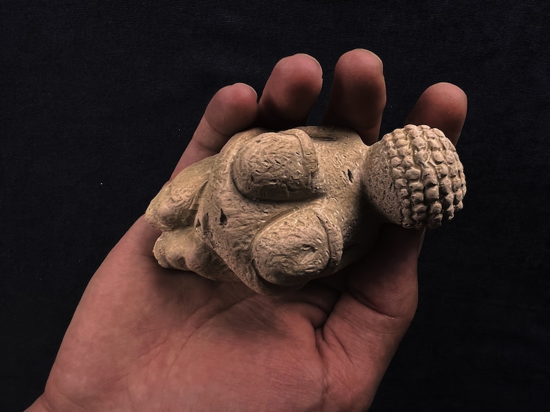 May include: A beige, abstract sculpture with a textured surface. The sculpture has rounded shapes and a detailed, patterned sphere at one end. The object is held in a hand against a dark background.
