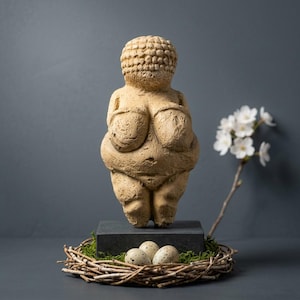 May include: A beige Venus figurine stands on a dark gray block, with a nest of twigs and speckled eggs in front. White flowers on a branch are to the right. The figurine has exaggerated features and a textured surface.