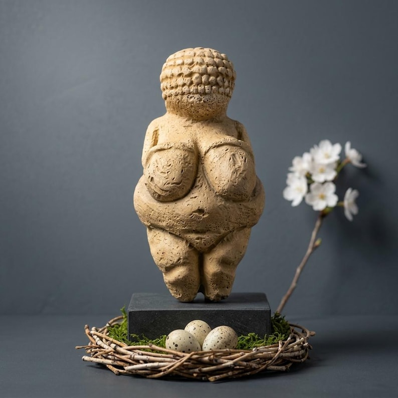 May include: A beige Venus figurine stands on a dark gray block, with a nest of twigs and speckled eggs in front. White flowers on a branch are to the right. The figurine has exaggerated features and a textured surface.