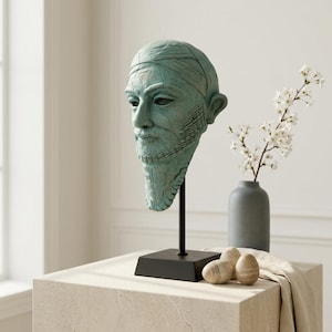 Puede incluir: Un busto de color turquesa de un hombre con barba y pañuelo en la cabeza, montado sobre un soporte negro. La escultura se exhibe sobre un bloque de piedra de color claro, con un jarrón de flores blancas y piedras decorativas cerca. El busto tiene una apariencia desgastada.