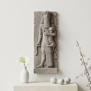 Puede incluir: Un relieve mural rectangular gris representa una figura barbuda que sostiene un león. La figura lleva una túnica larga y sostiene un objeto en forma de serpiente. Un jarrón blanco con un lirio y esferas de mármol están en un estante debajo.