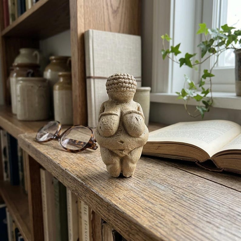 May include: A small, beige Venus figurine with exaggerated breasts and a detailed hairstyle. The sculpture is displayed on a wooden shelf with books, eyeglasses, and a window with greenery in the background. The figurine is a representation of prehistoric art.
