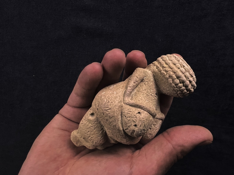 May include: A small, tan-colored figurine with a textured surface, held in a hand. The figurine has a detailed head with a woven pattern and a stylized body. The background is a solid black.