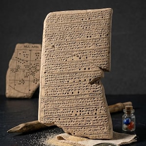 May include: A fragment of a clay tablet with cuneiform script, possibly ancient Mesopotamian. The tablet is a light brown color and has a broken edge. Other artifacts include a small map, a wooden stylus, and a small glass bottle with colored marbles.