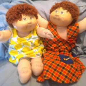 May include: Two Cabbage Patch Kids dolls, one wearing a yellow dress with a lemon print and the other wearing a red and orange plaid dress with a blue pocket.