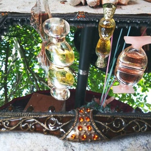 Pode incluir: Três frascos de perfume de vidro com tampas douradas e prateadas, refletidos em um espelho com uma moldura dourada ornamentada. Os frascos estão cheios de líquidos transparentes e coloridos.