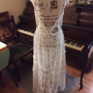 May include: A white lace maxi dress with a low back and a tiered skirt. The dress is on a mannequin and is displayed in front of a piano.