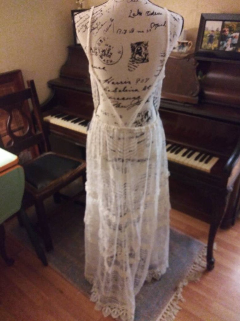 May include: A white lace maxi dress with a low back and a tiered skirt. The dress is on a mannequin and is displayed in front of a piano.