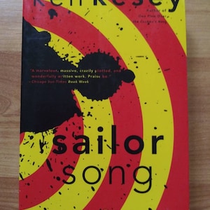 May include: A book cover with a red and yellow circular pattern. The title "sailor song" is in black text. The author's name "kenkesey" is at the top of the cover. The author is also identified as the author of "One Flew Over the Cuckoo's Nest". A quote from the Chicago Sun-Times Book Week is included: "A marvelous, massive, crazily plotted, and wonderfully written work. Praise be."
