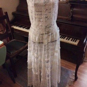 May include: A white lace dress with a sleeveless design and a flowing skirt. The dress is displayed on a mannequin in front of a vintage piano.
