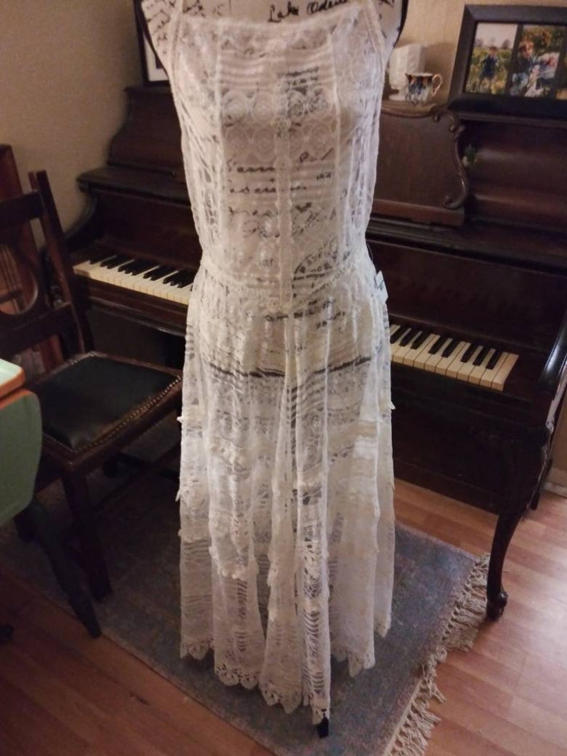May include: A white lace dress with a sleeveless design and a flowing skirt. The dress is displayed on a mannequin in front of a vintage piano.