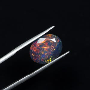 May include: An oval-shaped black opal gemstone featuring a faceted surface with vibrant flashes of red, orange, and yellow color play. The dark stone is held by a pair of silver-toned metal tweezers against a solid black background.