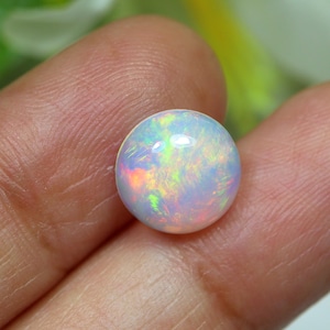 May include: A round, iridescent opal gemstone. The opal displays a milky white base color with flashes of green, yellow, and orange. The gemstone is smooth and polished, reflecting light.