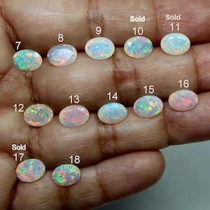 May include: A collection of oval-shaped, faceted opals arranged in rows, each labeled with a number from 7 to 18. The gemstones display iridescent flashes of green, pink, and blue against a translucent white base. Items 10, 11, and 17 are marked Sold.
