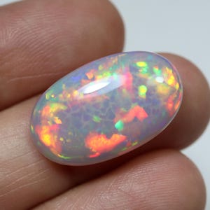 May include: An oval-shaped cabochon opal gemstone featuring a translucent milky white base with vibrant flashes of red, orange, yellow, and green play-of-color throughout the stone. The surface is smooth and polished with a domed top profile.