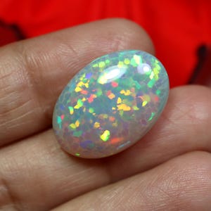May include: An oval-shaped opal gemstone with a white base and iridescent flashes of color. The gemstone displays a mosaic-like pattern with vibrant hues of green, yellow, and red. The opal is held in a hand, showcasing its polished surface.