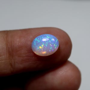 May include: An oval-shaped cabochon opal gemstone featuring a translucent white base with vibrant flashes of blue, green, and orange light. The stone has a smooth, polished surface that reflects light, showcasing a play-of-color effect throughout.