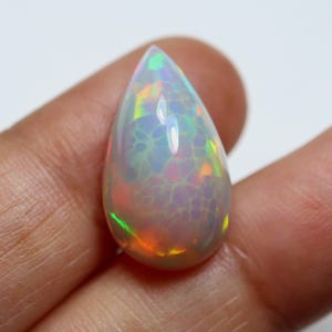 Könnte beinhalten: Ein tropfenförmiger Opal-Edelstein mit weißer Basis und innerem Farbspiel. Der Edelstein zeigt Blitze von Grün, Orange und Gelb. Die Oberfläche hat ein Rissmuster. Der Opal wird in einer Hand gehalten.