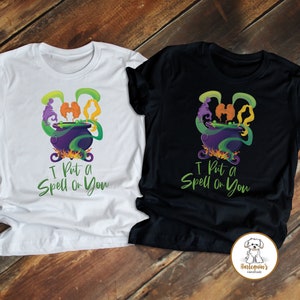 Pode incluir: Duas camisetas com tema de Halloween. Uma é branca e a outra é preta. Ambas apresentam um caldeirão com fumaça verde e o texto "I Put a Spell On You".