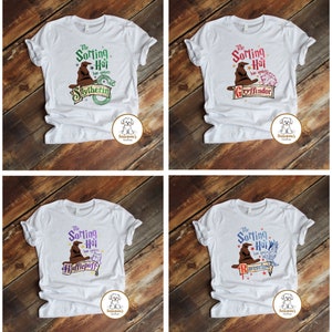 May include: Four white t-shirts with a graphic print of the Sorting Hat from the Harry Potter series. Each shirt features a different Hogwarts house crest: Slytherin, Gryffindor, Hufflepuff, and Ravenclaw.