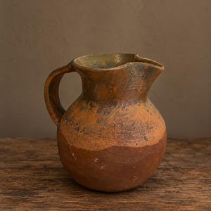 May include: A rustic, round ceramic pitcher with a wide handle and a pronounced pouring spout. The vessel features a two-tone finish, with a dark, matte reddish-brown base transitioning into a mottled, weathered ochre and charcoal upper section.