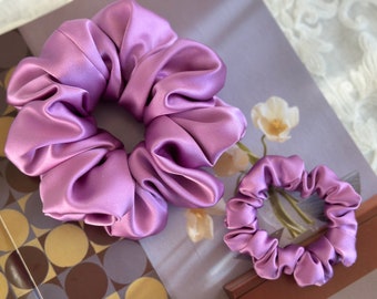 May include: Two purple satin scrunchies, one large and one small, on a patterned surface.