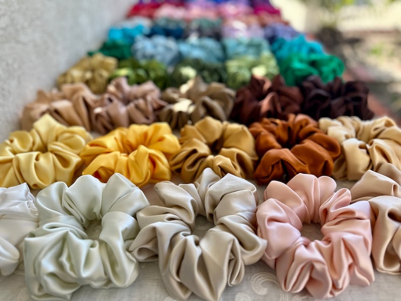 May include: A collection of colourful satin scrunchies in various shades of brown, yellow, beige, and pink. The scrunchies are arranged in a row, with the lighter colours at the front and the darker colours at the back.