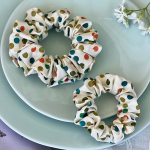 May include: Two white fabric scrunchies with green, orange, and blue polka dots. The scrunchies are on a light blue plate.
