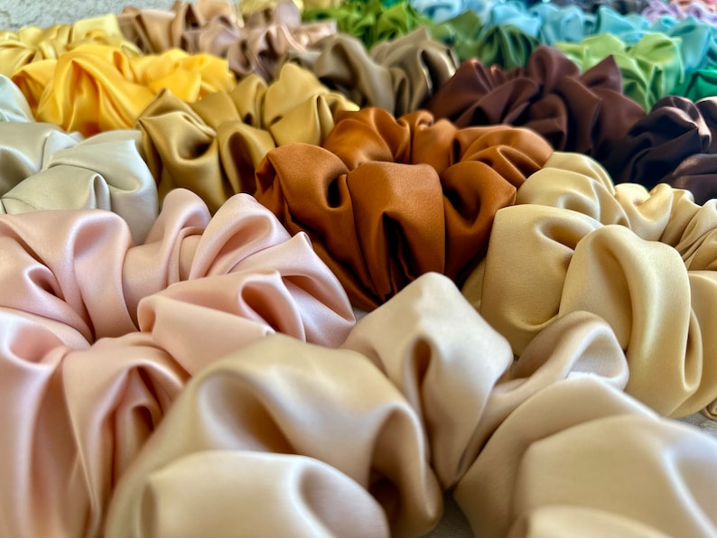 May include: A collection of colourful satin scrunchies in various shades of brown, beige, and pink. The scrunchies are arranged in a pile, showcasing their soft and luxurious texture.