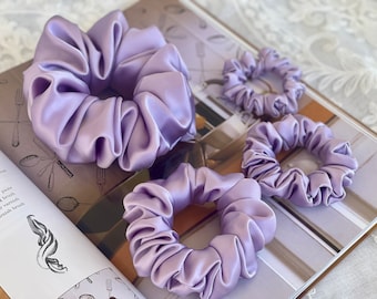 May include: Three lilac-colored satin scrunchies of different sizes. The scrunchies are made of soft, silky fabric and are perfect for holding hair in place.