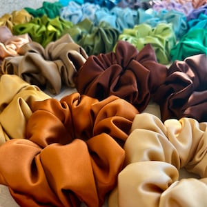 May include: A collection of colourful satin scrunchies in various shades of brown, beige, and green. The scrunchies are made of soft, silky fabric and are perfect for adding a touch of style to any hairstyle.