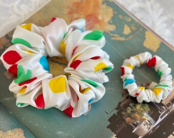 May include: Two white scrunchies with colorful polka dots. The larger scrunchie has red, yellow, blue, and green dots. The smaller scrunchie has red, yellow, and blue dots.