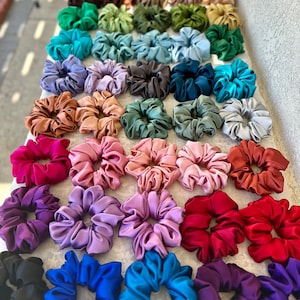 May include: A collection of colourful satin scrunchies in various shades of blue, purple, pink, red, green, brown, and black. The scrunchies are arranged in rows on a white surface.