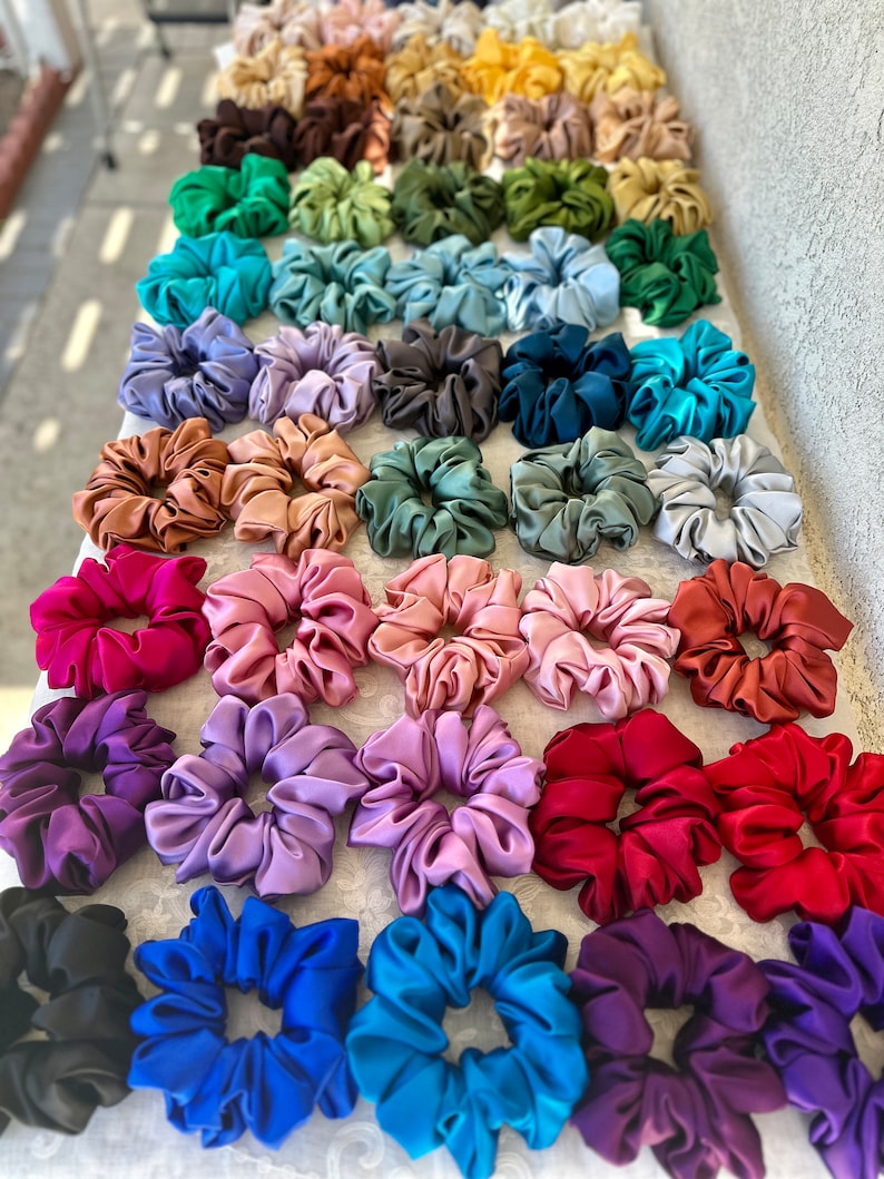 May include: A collection of colourful satin scrunchies in various shades of blue, purple, pink, red, green, brown, and black. The scrunchies are arranged in rows on a white surface.
