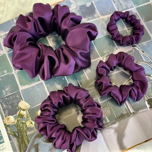May include: Four plum-coloured satin scrunchies of varying sizes are arranged on a tiled surface. The scrunchies are bunched and gathered, with a shiny, smooth texture. A small bouquet of white flowers is visible in the background.