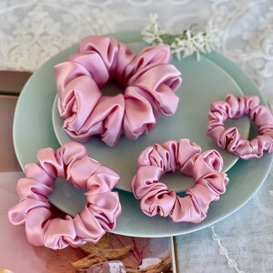 May include: Four pink satin scrunchies of different sizes on a light blue plate. The scrunchies are arranged in a circle with the largest scrunchie in the center.