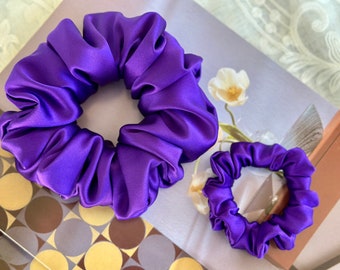 May include: Two purple satin scrunchies, one large and one small. The scrunchies are made of soft, silky fabric and are perfect for holding hair in place.