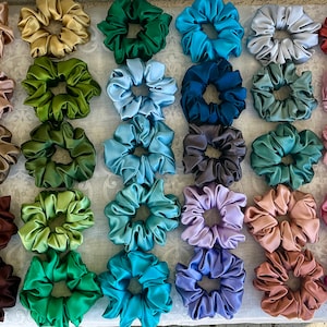 May include: A collection of 40 colourful satin scrunchies in various shades of green, blue, purple, brown, red, and white. The scrunchies are arranged in rows on a white surface.